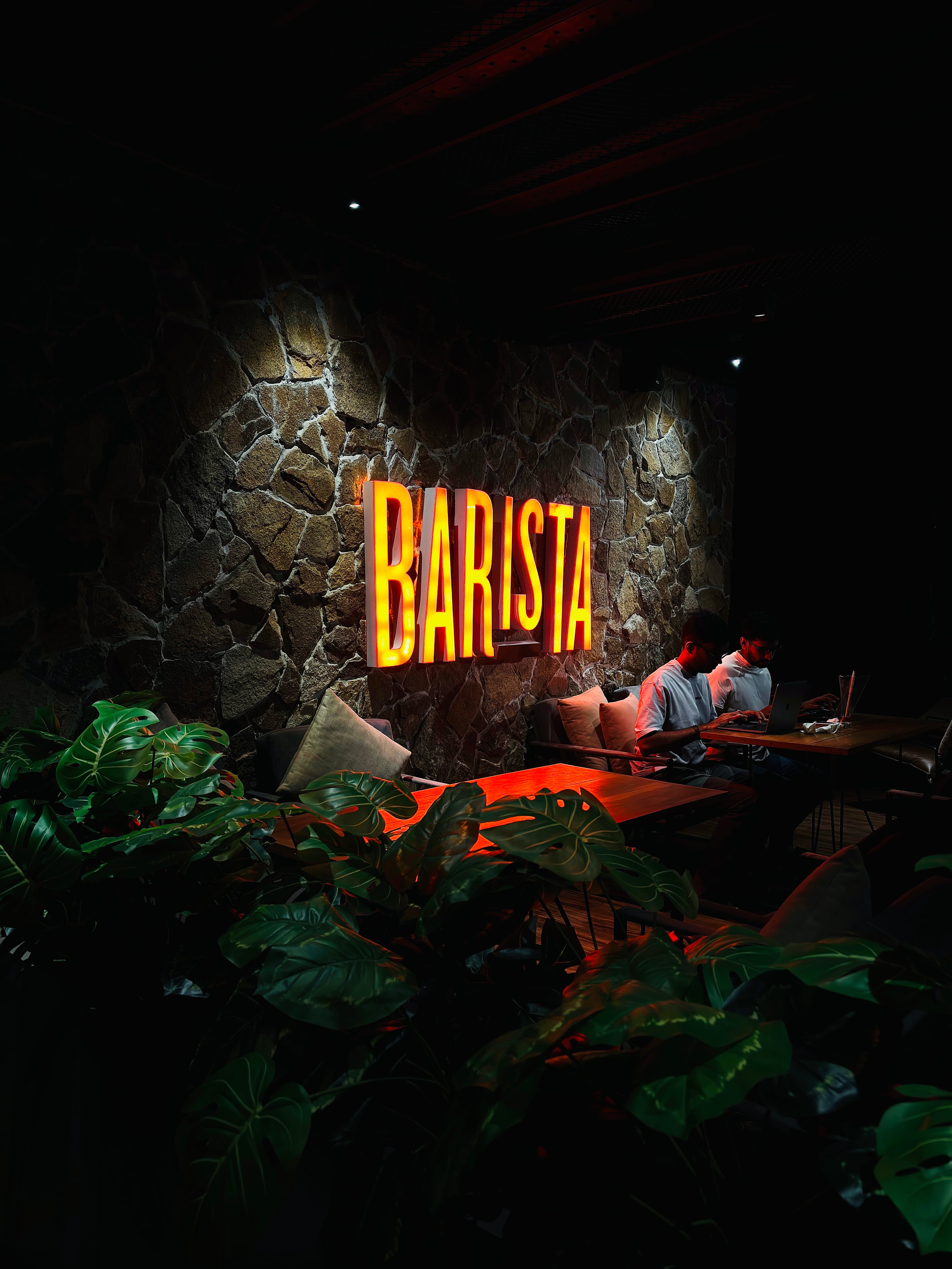 Neon 'BARISTA' sign glowing on a dark stone wall in a dimly lit cafe with lush plants in the foreground