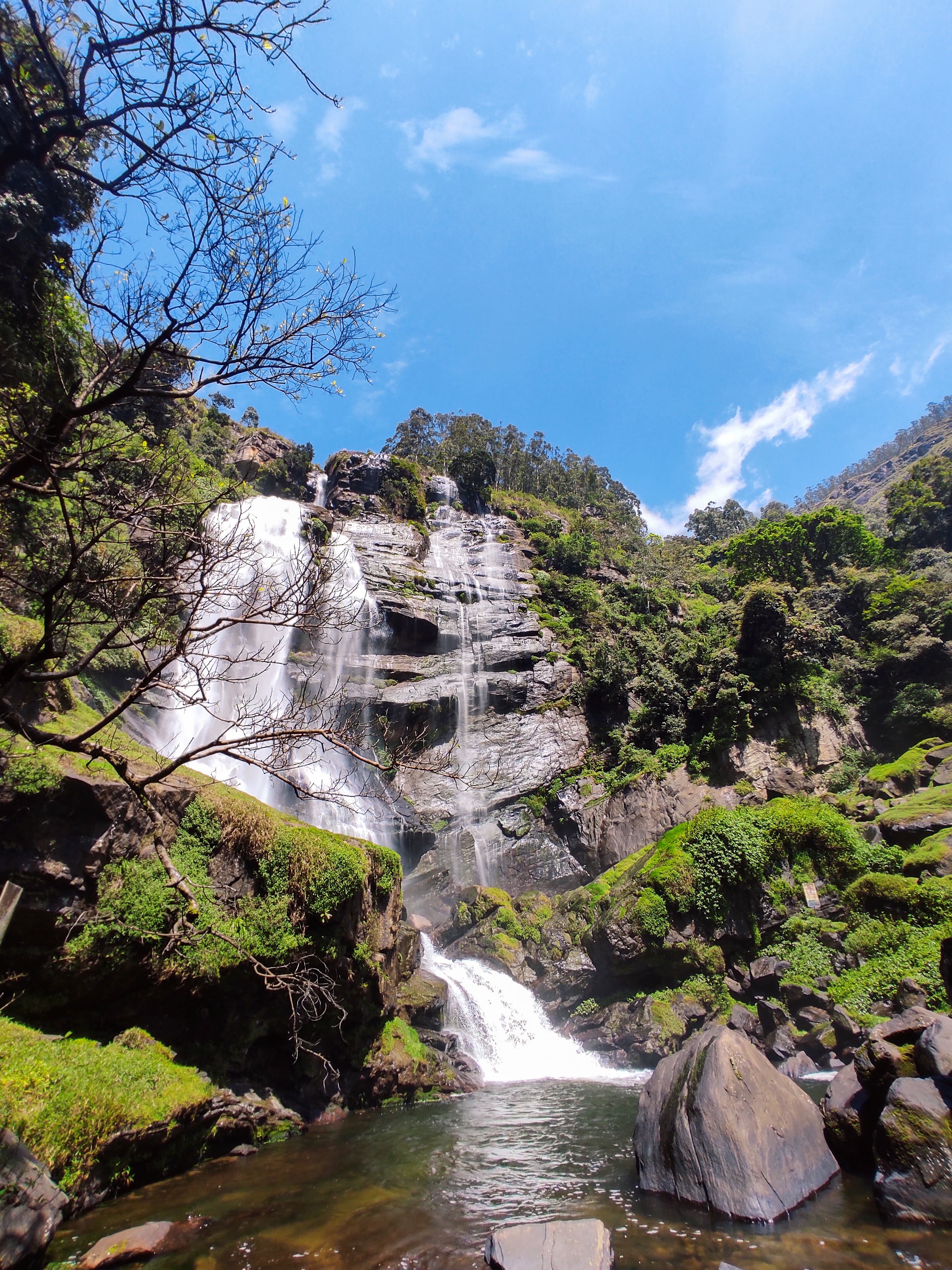 The wide, cascading Bomburu Ella waterfall flowing over mossy rocks on a bright sunny day with blue skies