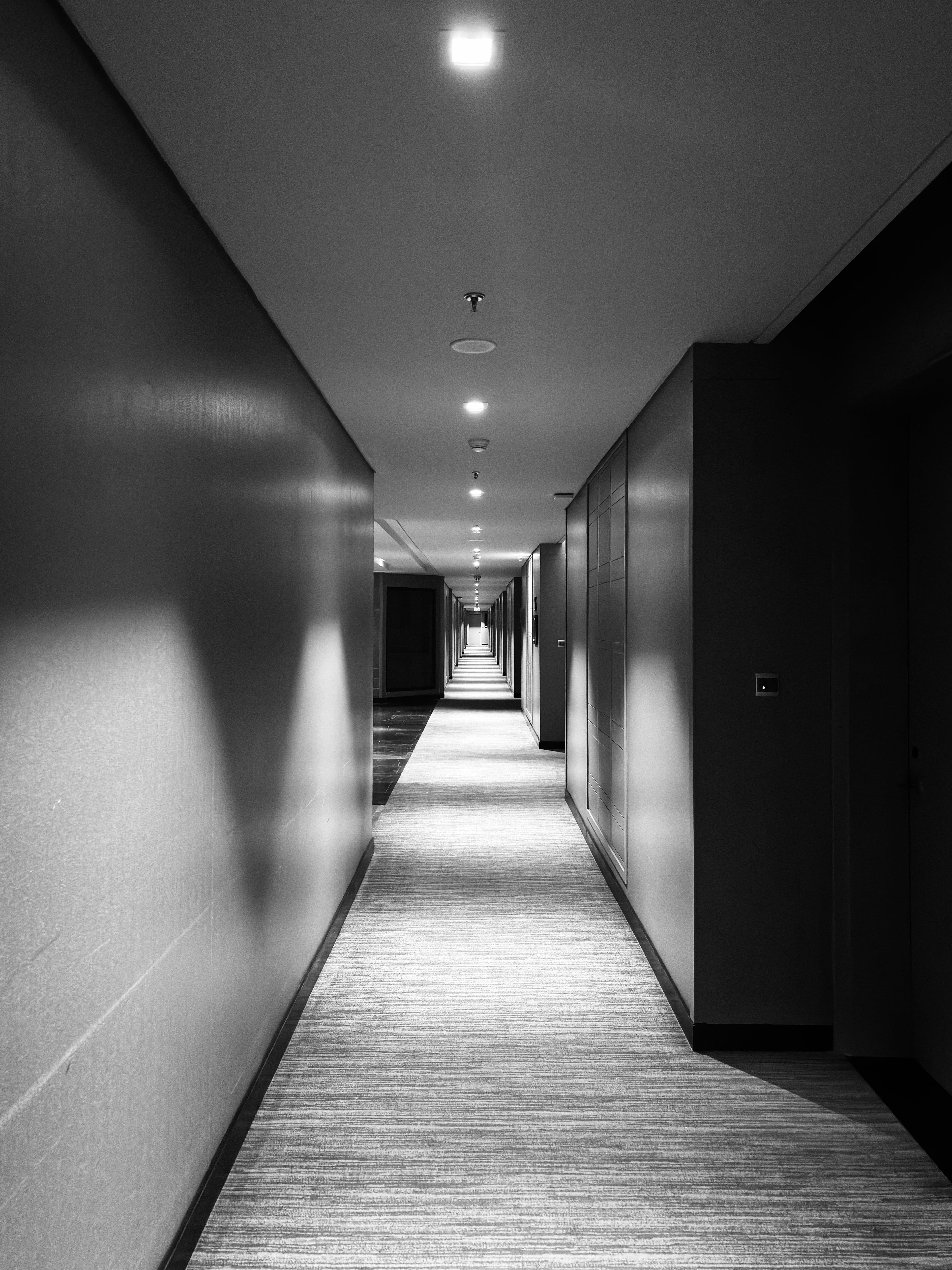 A black and white photograph of a long, illuminated, empty hotel corridor