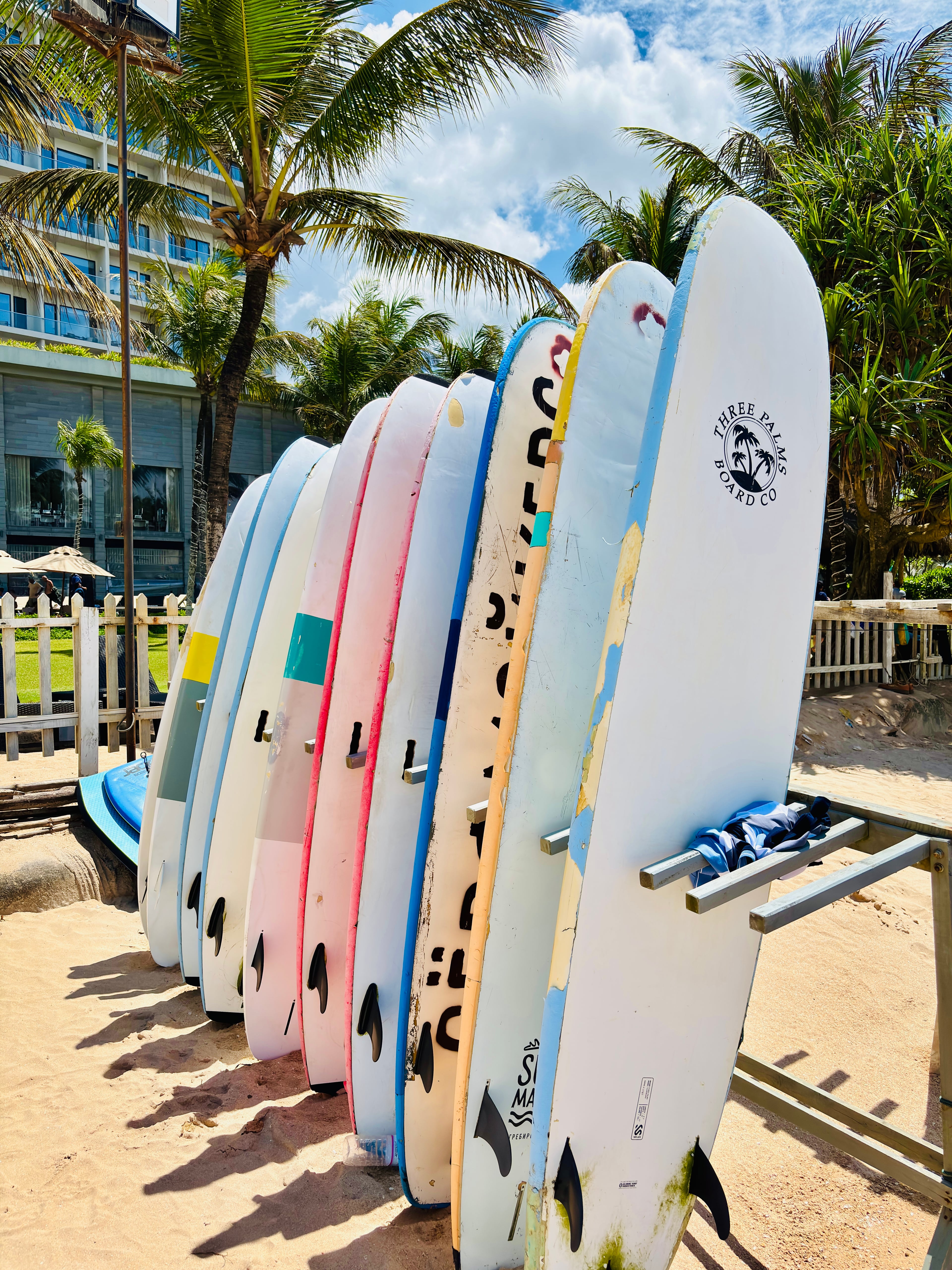 A row of colorful surfboards standing upright on a rack in the sand, with palm trees and a resort in the background