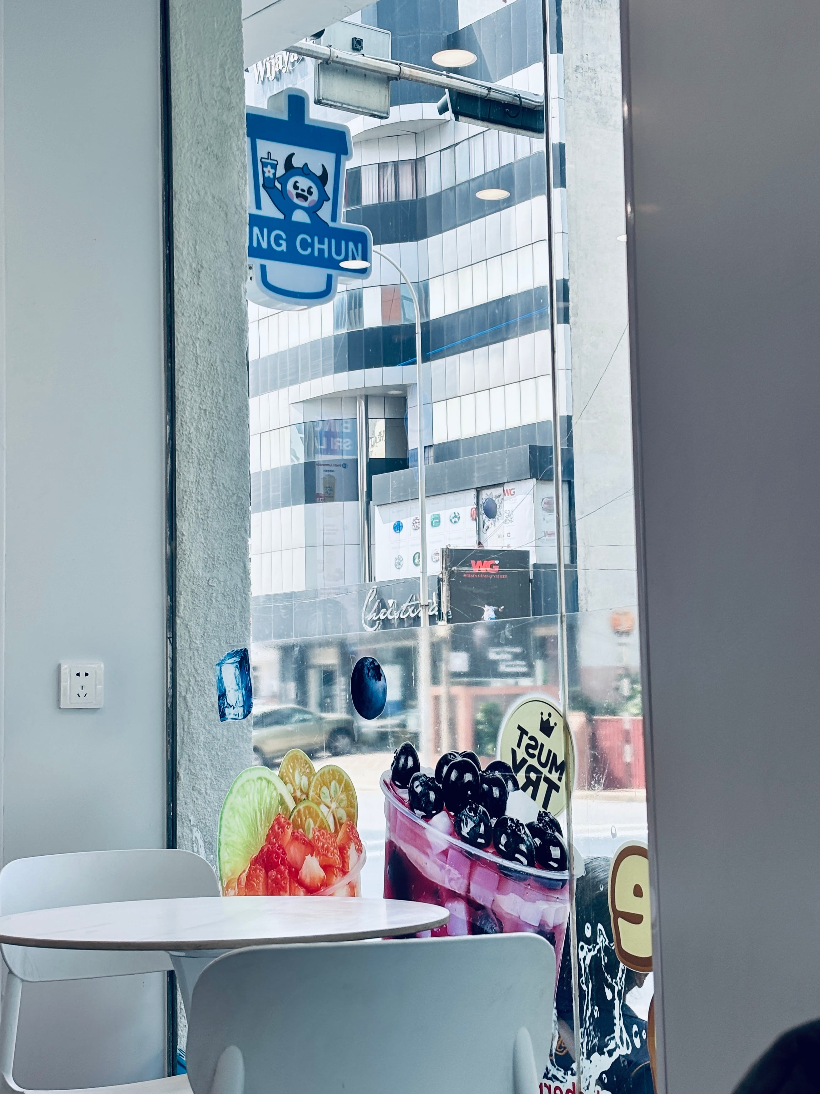 View through a cafe window with fruit drink decals, showing a street and a modern glass building outside