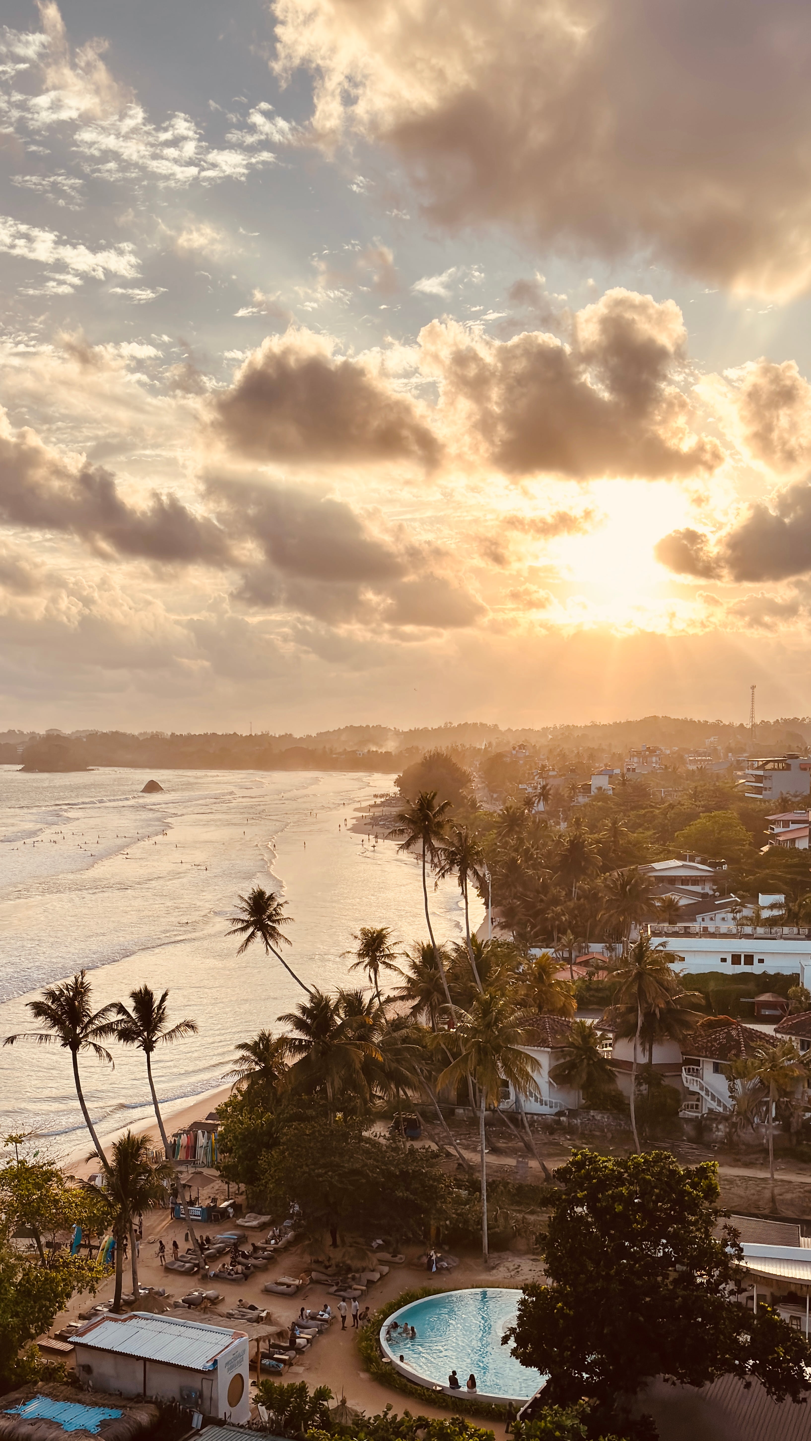 A stunning golden sunset casting warm light over a tropical coastline with palm trees, a circular pool, and ocean waves