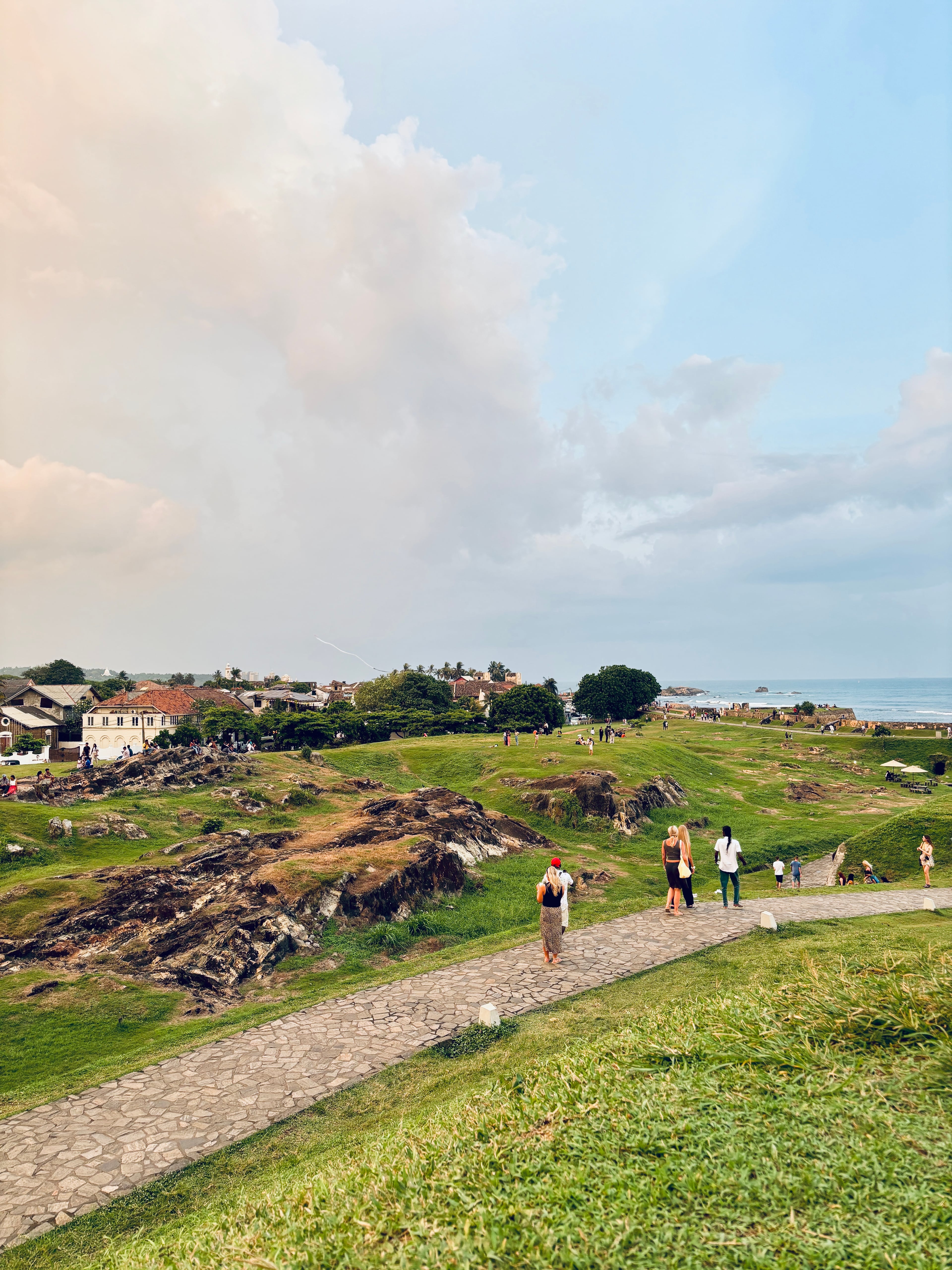 People walking along a stone path on grassy ramparts overlooking a historic coastal town and the ocean