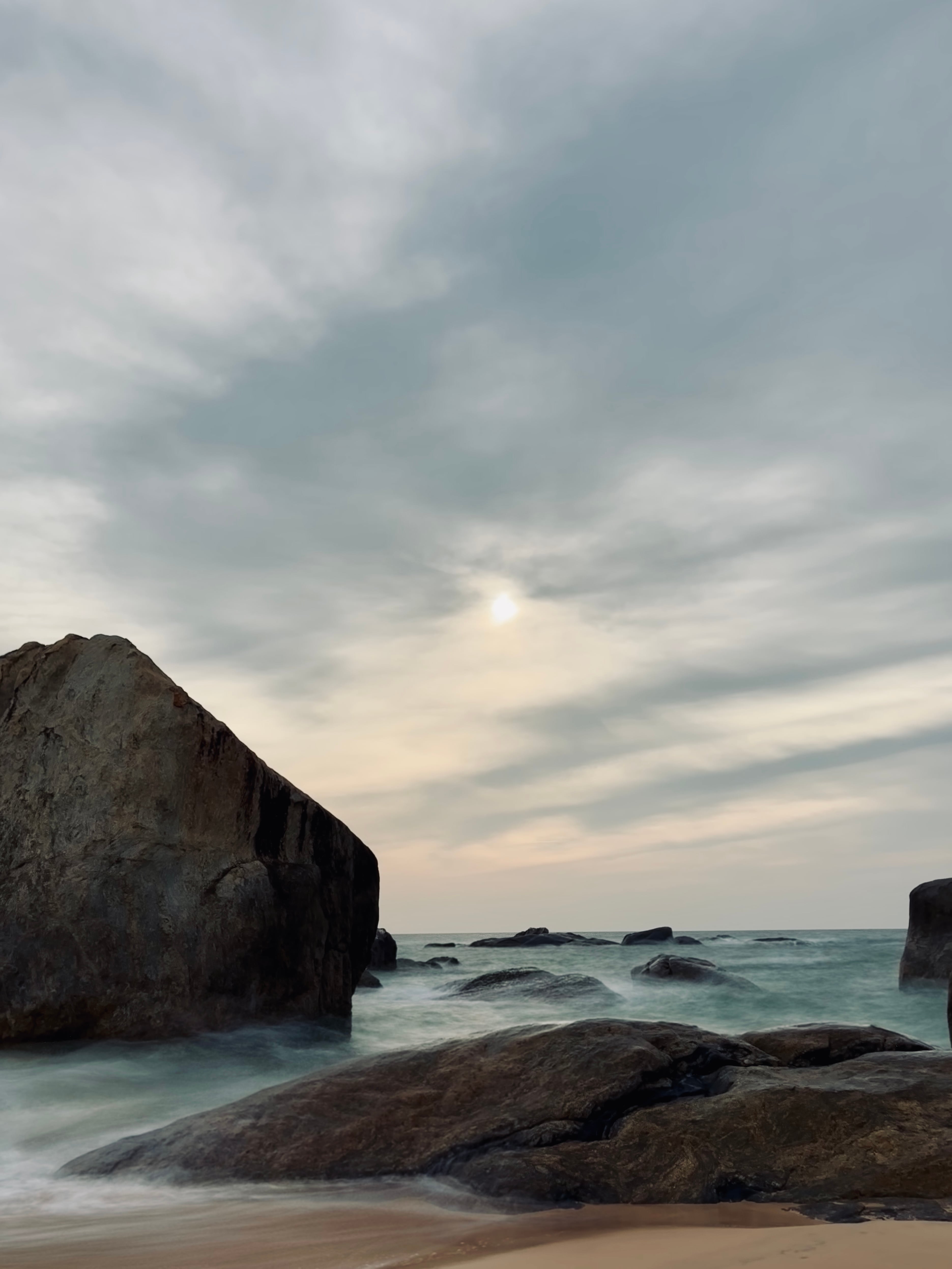 Large, dark boulders on a sandy beach with ocean water smoothed into mist by a long exposure under a cloudy sky
