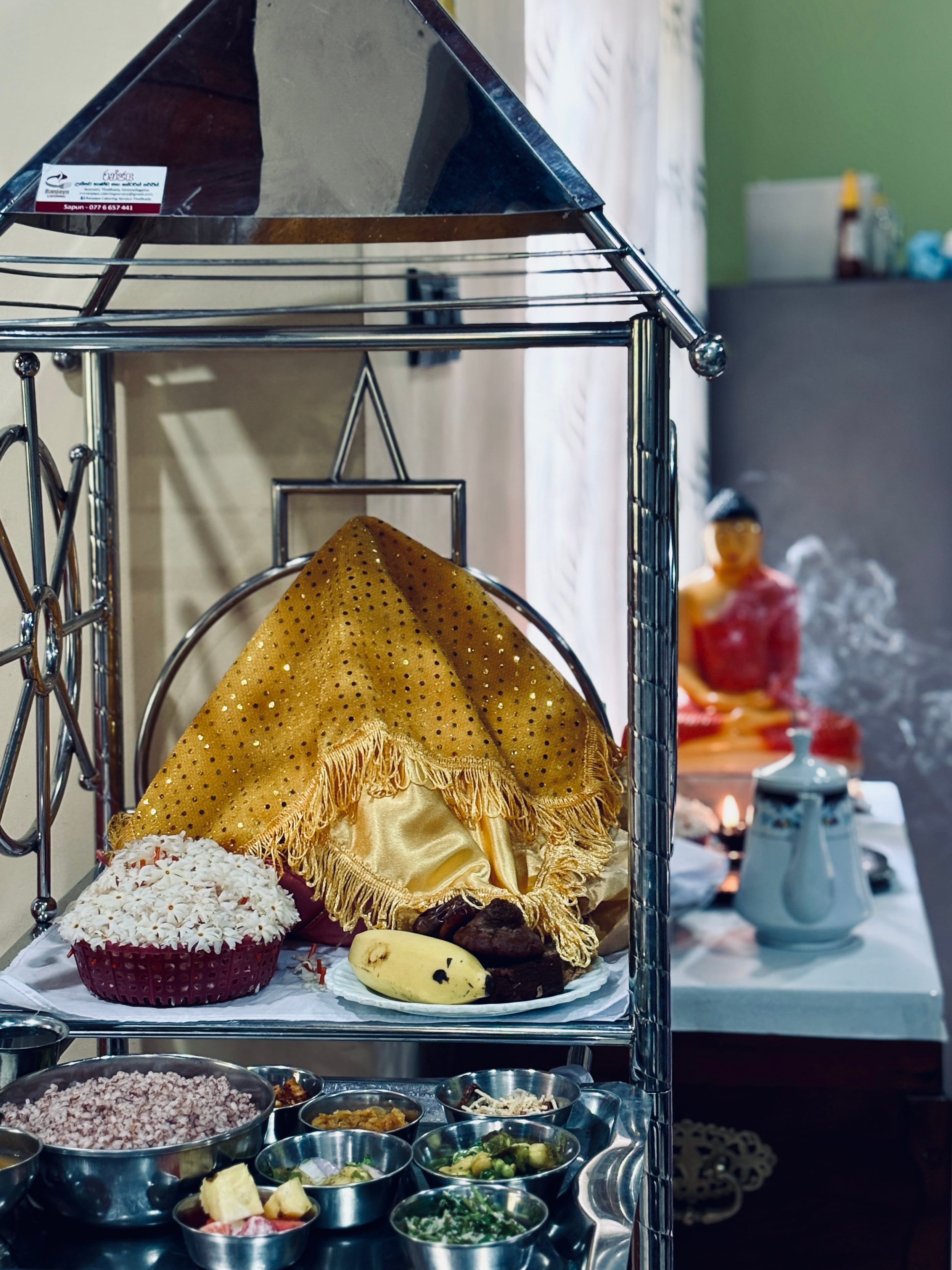 A traditional Sri Lankan alms giving setup with curries in metal bowls and a covered offering on a silver stand, with a blurred Buddha statue in the background