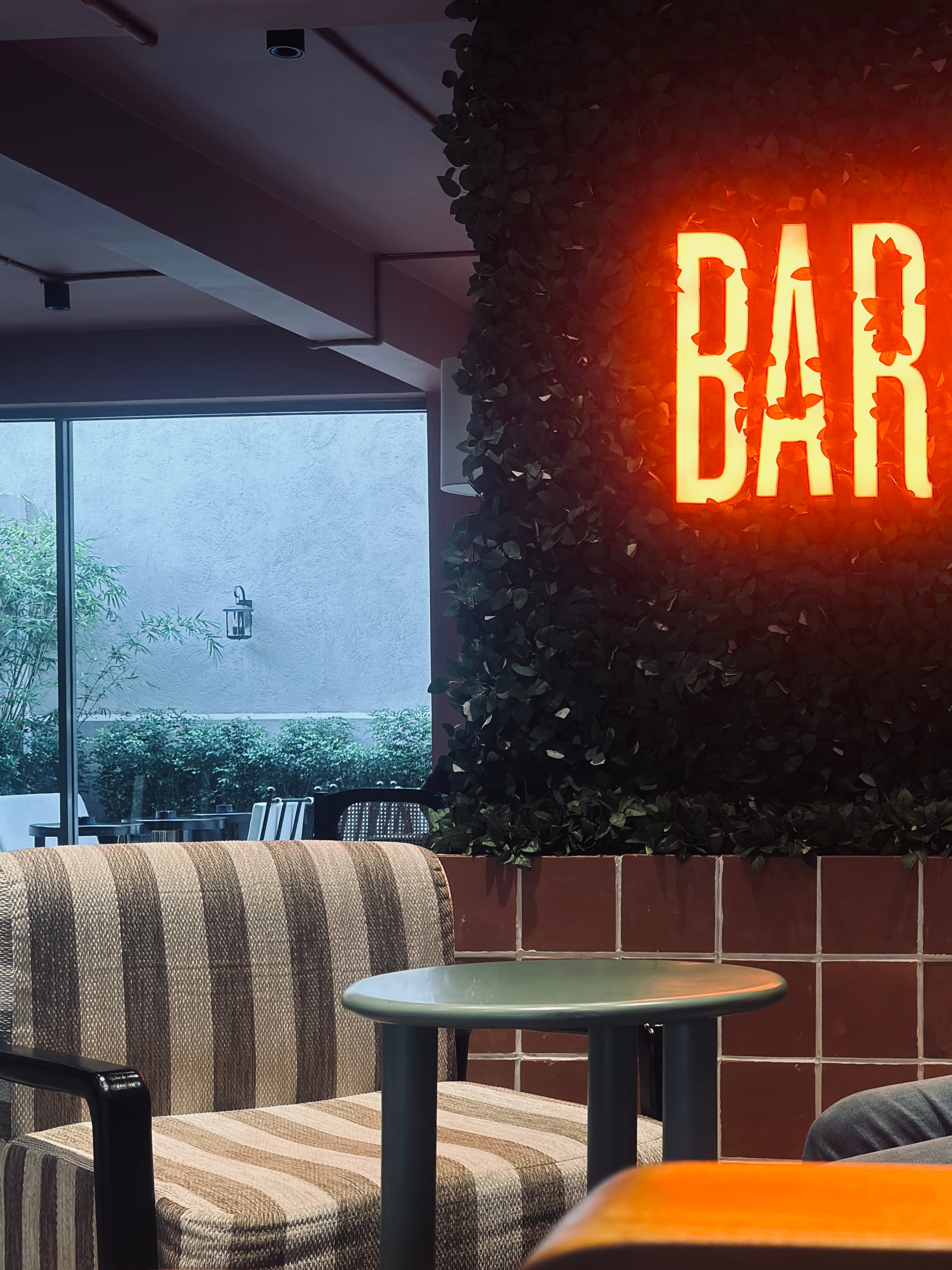 A glowing orange neon sign mounted on a wall covered in green leaves inside a Barista cafe, with a striped armchair and table in the foreground