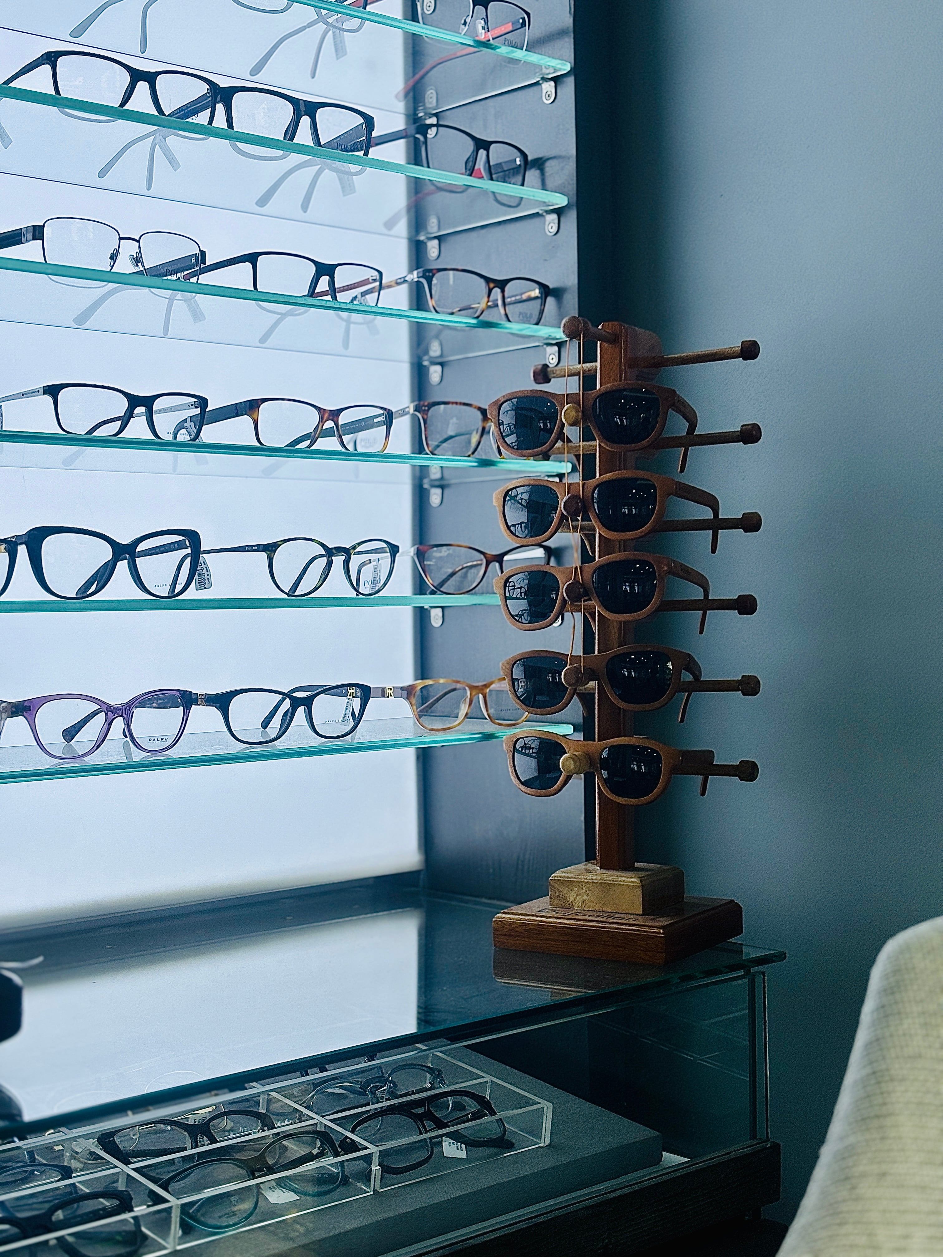 Rows of eyeglasses displayed on illuminated glass shelves, next to a wooden stand holding sunglasses with wooden frames