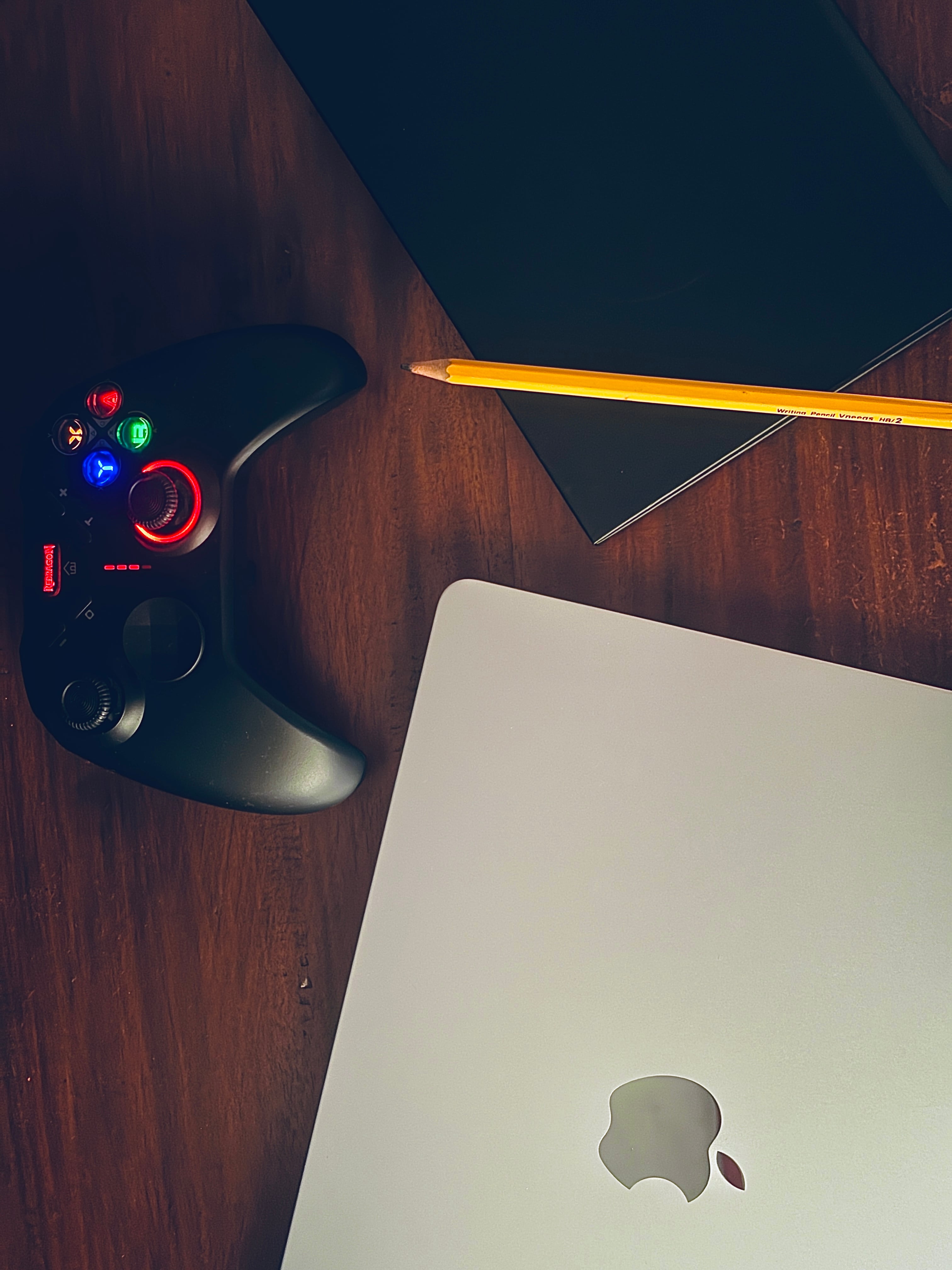 A top-down view of a silver MacBook, a black glowing game controller, a black notebook, and a yellow pencil on a dark wooden desk