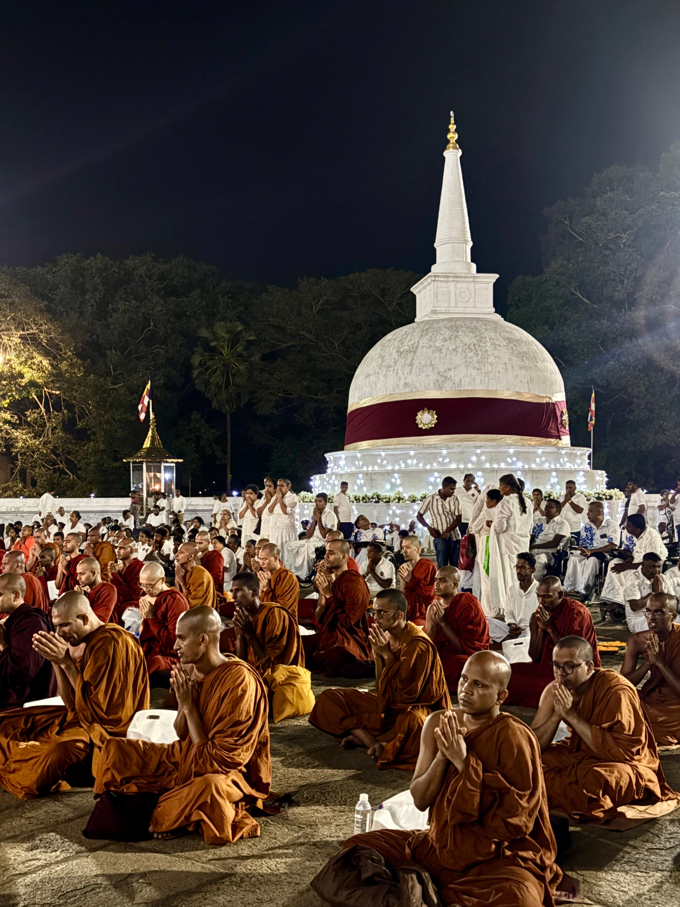 Dozens of Buddhist monks in orange robes seated in prayer before a brightly lit white stupa at night