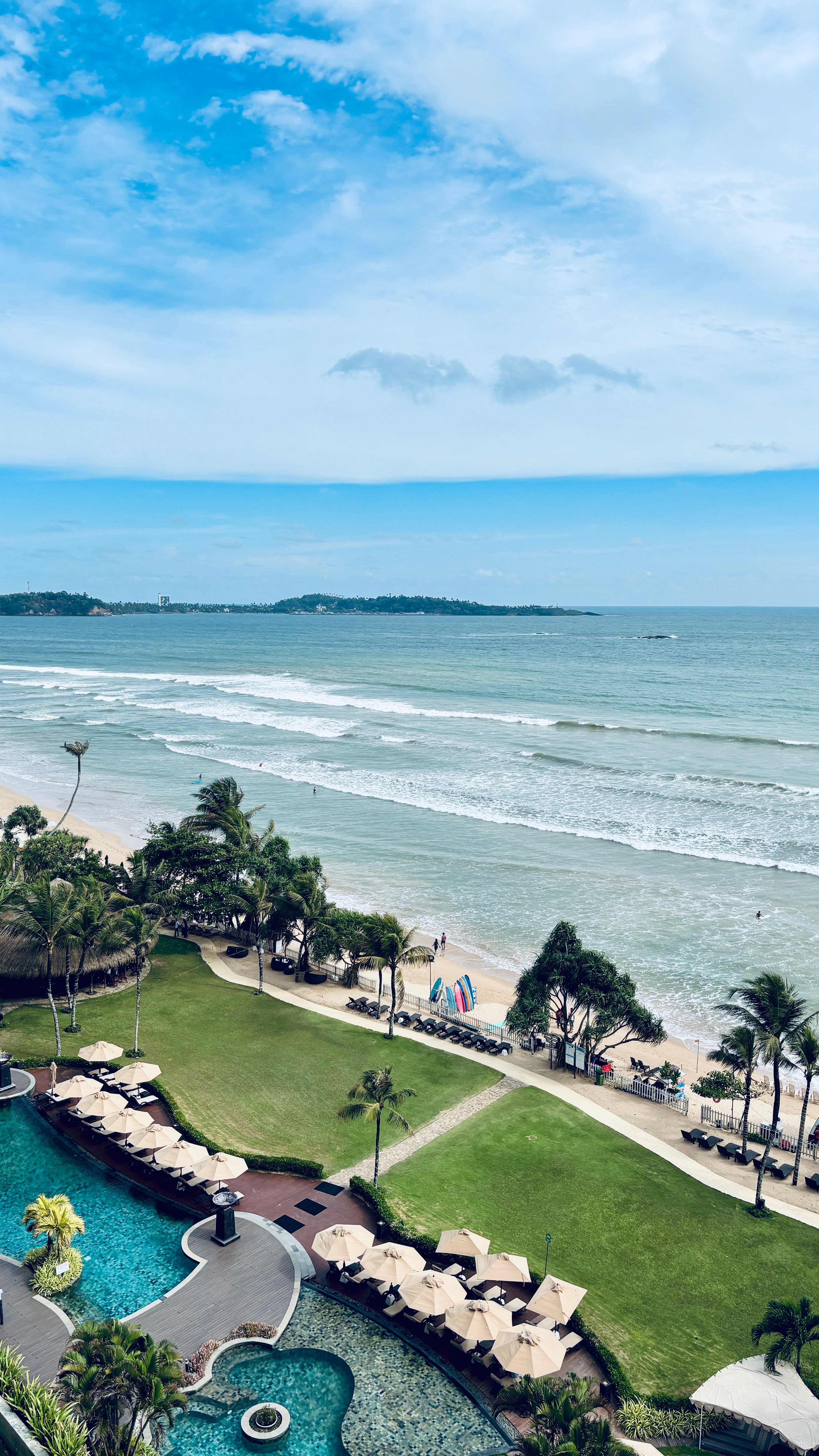 An elevated view of a coastal resort featuring a swimming pool, manicured lawns, palm trees, and a sandy beach facing the ocean