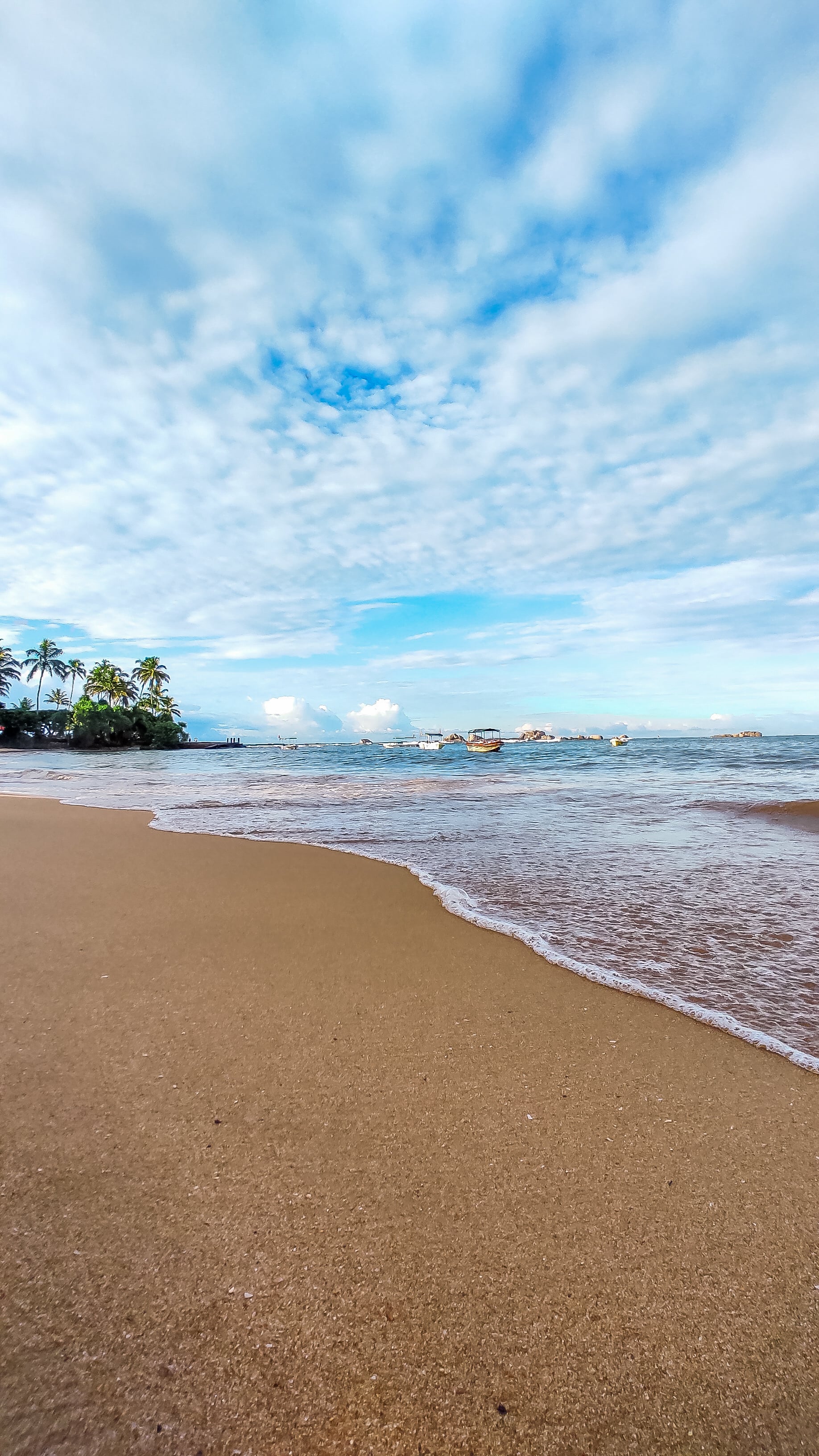 The serene shoreline of Hikkaduwa beach in the morning, with gentle waves and fishing boats in the distance under a partly cloudy sky.