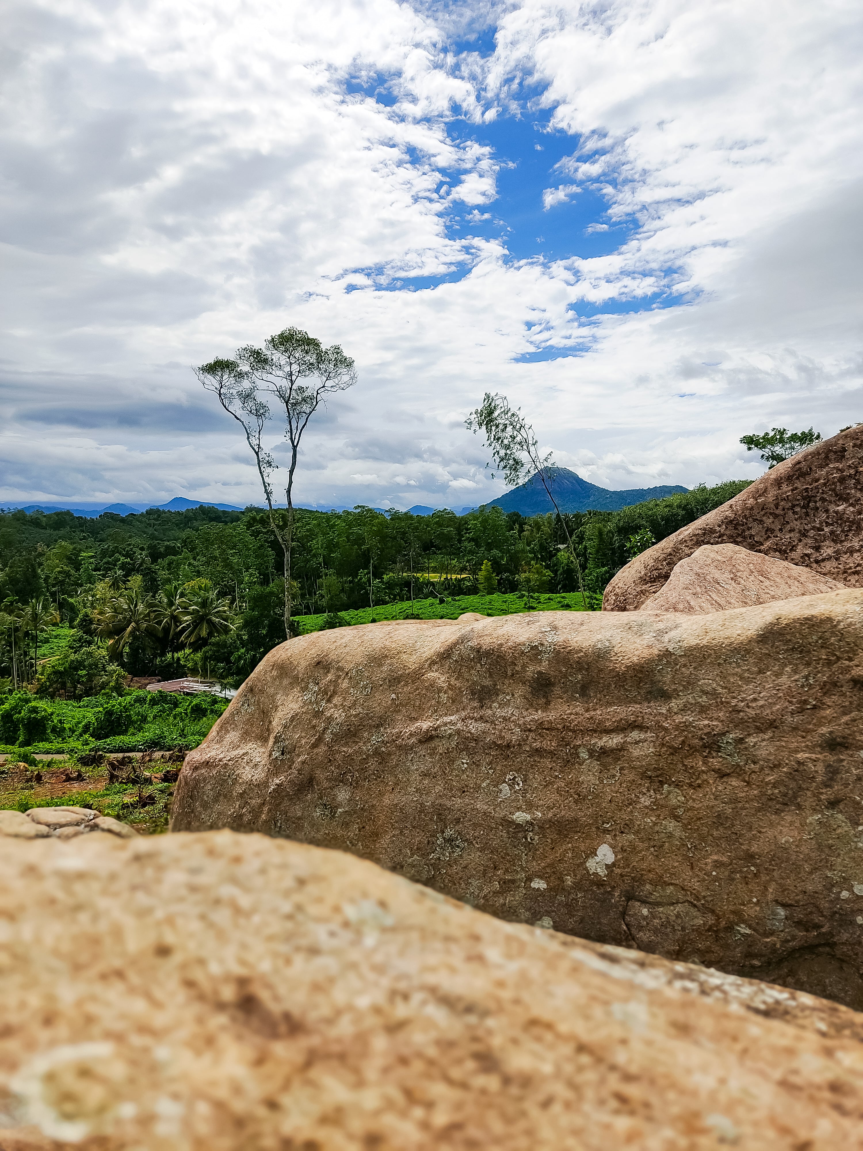 A scenic view of a green valley and distant mountains near Kandy, with large, weathered boulders in the foreground.