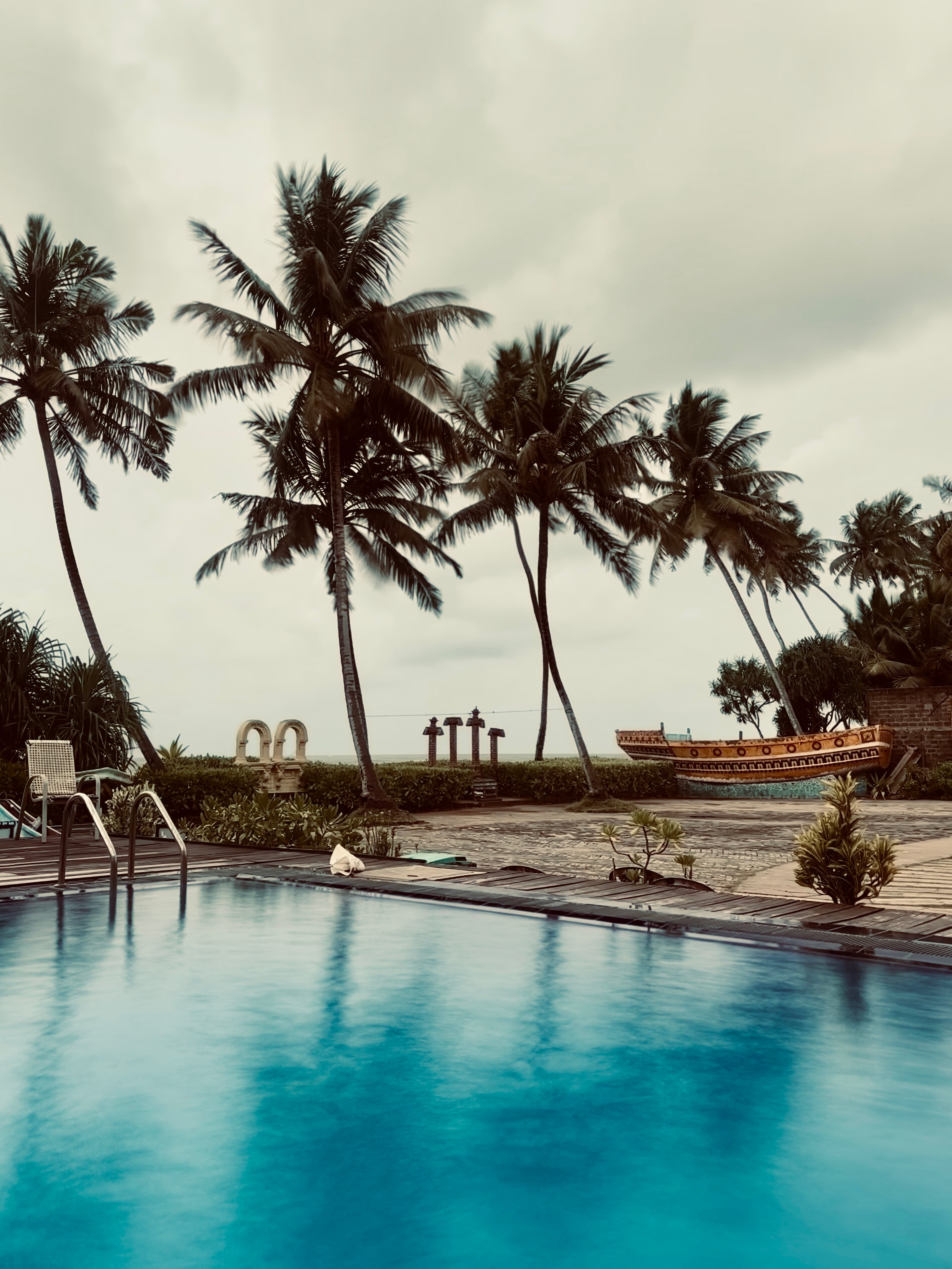 A swimming pool reflecting tall palm trees on an overcast day, with the ocean and a decorative boat in the background