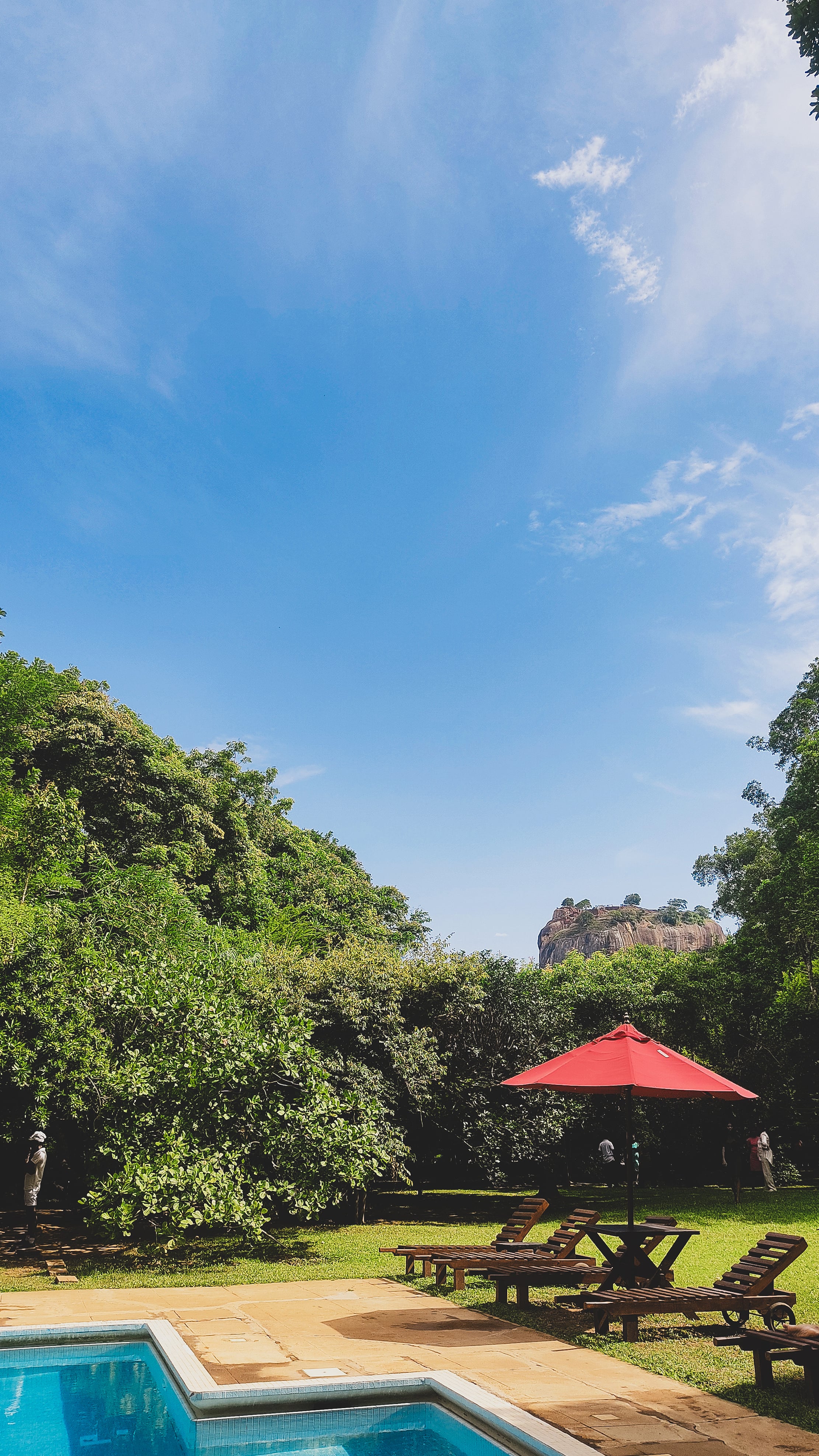 A view of the Sigiriya rock fortress in the distance from a resort with a swimming pool and red umbrella in the foreground